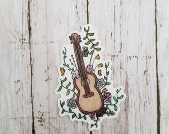 Op de afbeelding: Een sticker met een bruine akoestische gitaar met een bloemenontwerp. De gitaar is omringd door groene bladeren, roze, gele en witte bloemen en vlinders.