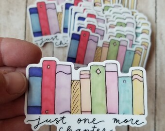 Peut inclure: Un autocollant blanc avec une pile de livres peints à l'aquarelle de couleurs vives et le texte "Just one more chapter".