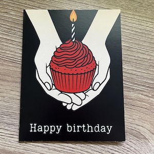 May include: A black birthday card with a red cupcake illustration. The cupcake has a lit candle and is held by two white hands. The words "Happy birthday" are printed in white at the bottom of the card.