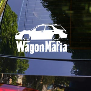 May include: A white decal of a station wagon with the text "Wagon Mafia" in a bold font is affixed to the rear window of a black car. The car is parked outdoors, with trees and other vehicles visible in the background.