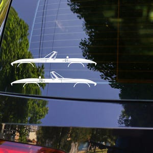 May include: A black car's rear window displays two white convertible car decals. The decals are side profiles of classic convertibles. The car is parked outdoors with trees visible in the background.