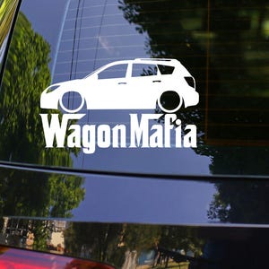 May include: A white vinyl decal of a station wagon with the text "Wagon Mafia" in a bold, sans-serif font. The decal is affixed to the rear window of a black car. The car is parked outdoors with trees in the background.