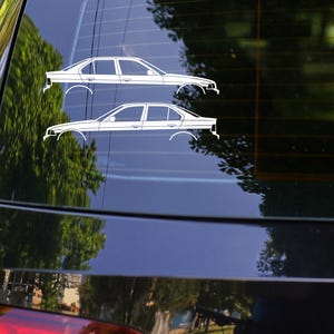 Pode incluir: Autocolantes brancos com contorno de carro no vidro traseiro de um carro preto. Os autocolantes retratam dois perfis laterais de um sedan. A luz traseira do carro &eacute; vermelha. O fundo mostra &aacute;rvores e outros carros.