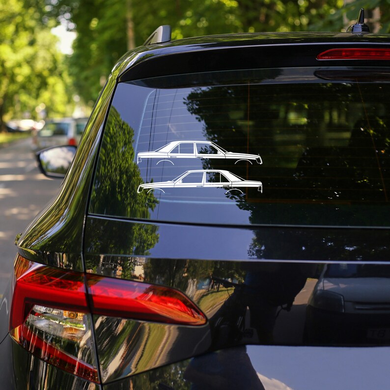 May include: Two white car decals are affixed to the rear window of a black vehicle. The decals depict side profiles of classic sedans. The background shows a blurred street scene with trees and other vehicles.