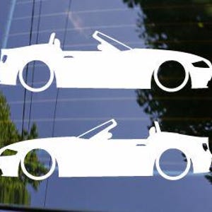 May include: Two white convertible car decals. The decals are side profiles of the car with the top down. The decals are on a window with a blurred background of trees and sky.