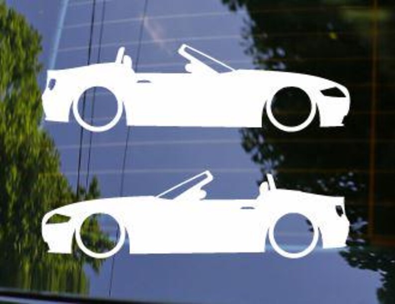 May include: Two white convertible car decals. The decals are side profiles of the car with the top down. The decals are on a window with a blurred background of trees and sky.