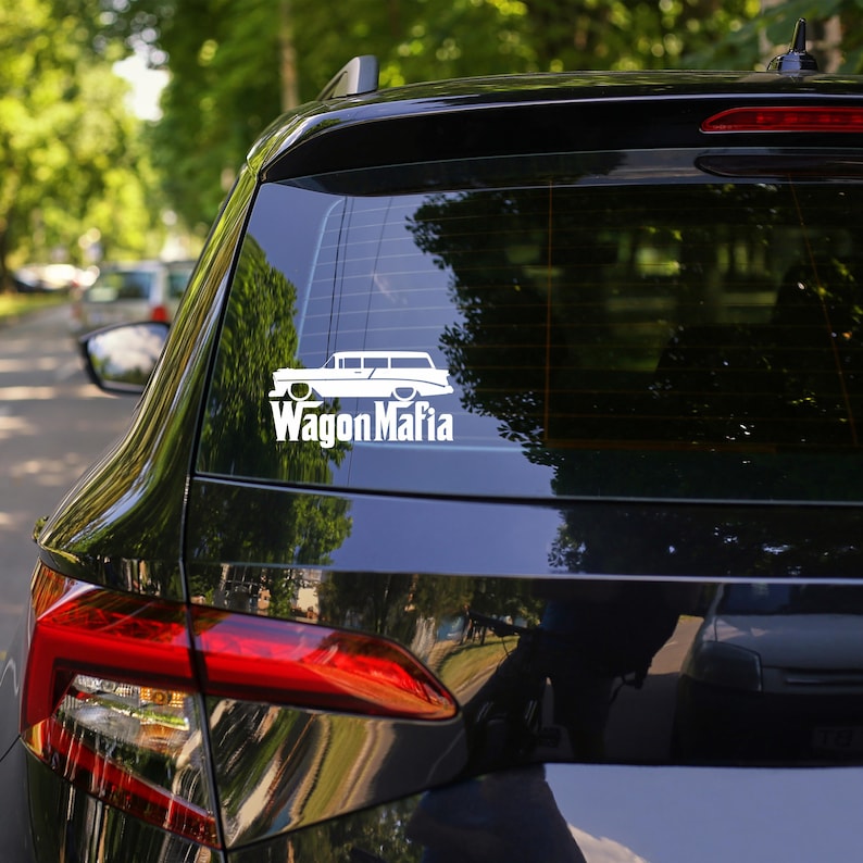 May include: A white sticker featuring a vintage estate car design with the words "Wagon Mafia". The decal is applied to the rear window of a black car. The background shows trees and a road, slightly out of focus.