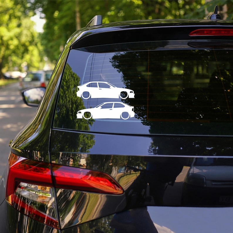 Puede incluir: Dos calcoman&iacute;as blancas de coche se muestran en la ventana trasera de un veh&iacute;culo negro. Las calcoman&iacute;as representan el perfil lateral de un coche, con detalles de las ruedas y la forma de la carrocer&iacute;a. El fondo muestra una vista borrosa de &aacute;rboles y otros coches.