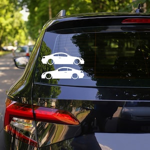 Könnte beinhalten: Zwei weiße Autoaufkleber sind auf der Heckscheibe eines schwarzen Fahrzeugs angebracht. Die Aufkleber zeigen stilisierte Seitenansichten eines Coupés mit kreisförmigen Raddesigns. Das Auto parkt im Freien, mit einem verschwommenen grünen Hintergrund.