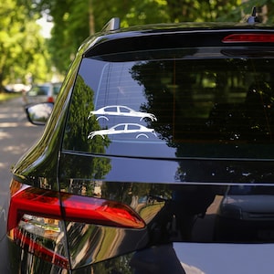 May include: Two white car decals are displayed on the rear window of a black car. The decals are minimalist outlines of saloon cars. The car is parked outside, with trees and other vehicles visible in the background, reflecting in the glass.