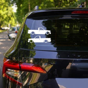 May include: Two white truck decals are affixed to the rear window of a black vehicle. The decals are identical, showing a side profile of a lowered pickup truck. The background shows a blurred view of trees and other vehicles.