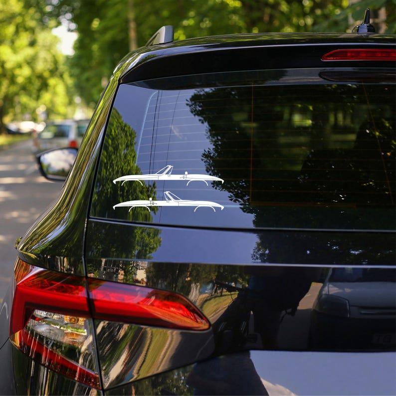 May include: A black car's rear window displays two white convertible car decals. The decals are side profiles of classic convertibles. The car is parked outdoors with trees visible in the background.