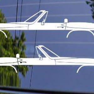 May include: Two white convertible car decals are displayed against a dark background. The decals are a minimalist design, featuring the car's outline with details like the windshield, wheels, and a small seat. The image is a close-up, highlighting the car's sleek shape.