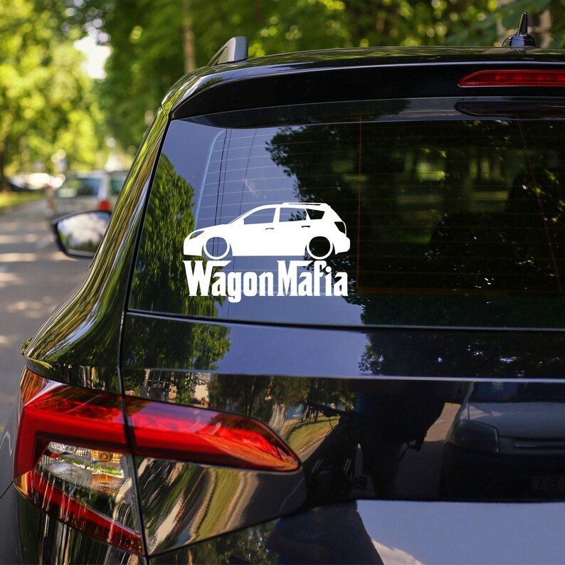 May include: A white vinyl decal of a station wagon with the text "Wagon Mafia" in a bold, sans-serif font. The decal is affixed to the rear window of a black car. The car is parked outdoors with trees in the background.