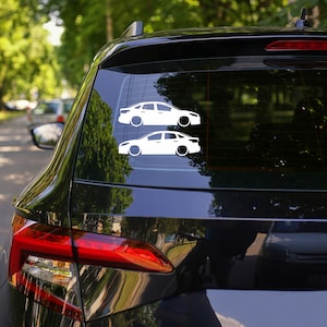 May include: Two white car stickers are displayed on the rear window of a black car. The stickers show side profiles of cars, featuring a low-slung design with black wheels. The background is a blurred view of trees and other vehicles.