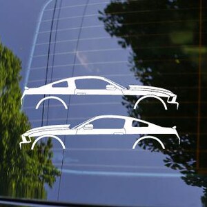 May include: Two white car decals of a classic sports car are displayed on a window. The decals are side profile silhouettes, showcasing the car's body lines and wheels. The background includes a blurred view of trees.