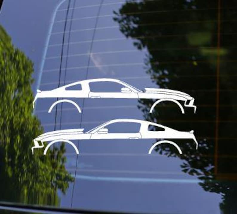 May include: Two white car decals of a classic sports car are displayed on a window. The decals are side profile silhouettes, showcasing the car's body lines and wheels. The background includes a blurred view of trees.