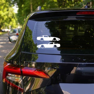May include: Two white car decals are affixed to the rear window of a black vehicle. The decals depict a side profile of a classic sports car. The car decals are a simple, minimalist design.