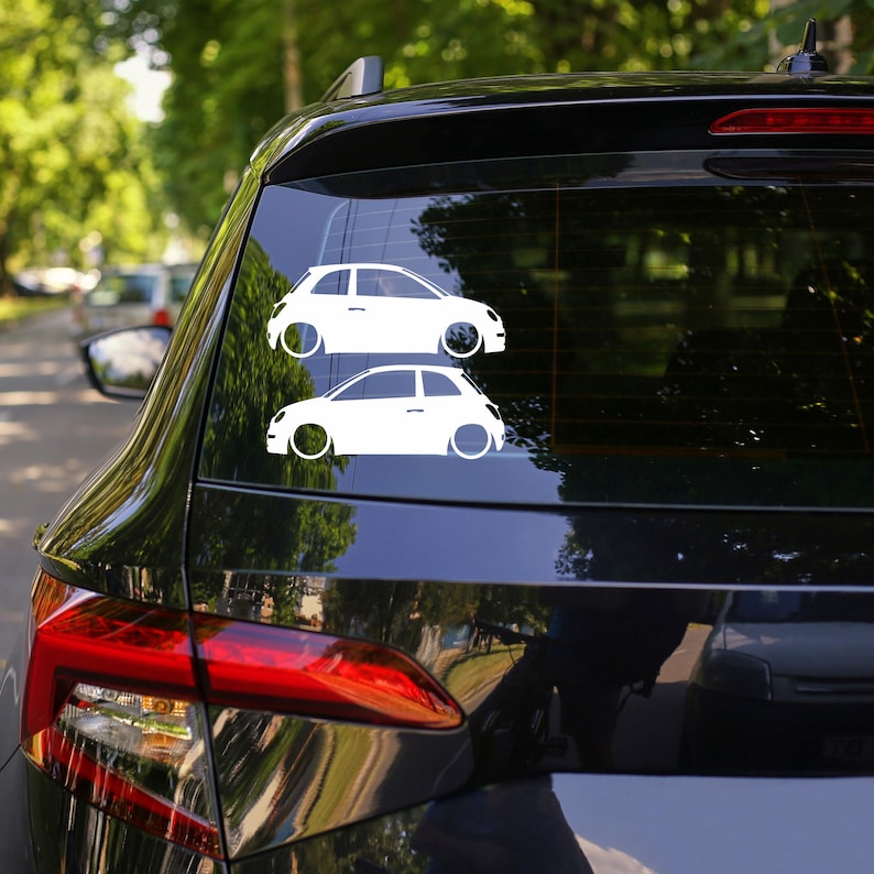 May include: Two white car decals are affixed to the rear window of a black vehicle. The decals depict stylized, side-view illustrations of small cars with large wheels. The background shows a blurred view of trees and other vehicles.