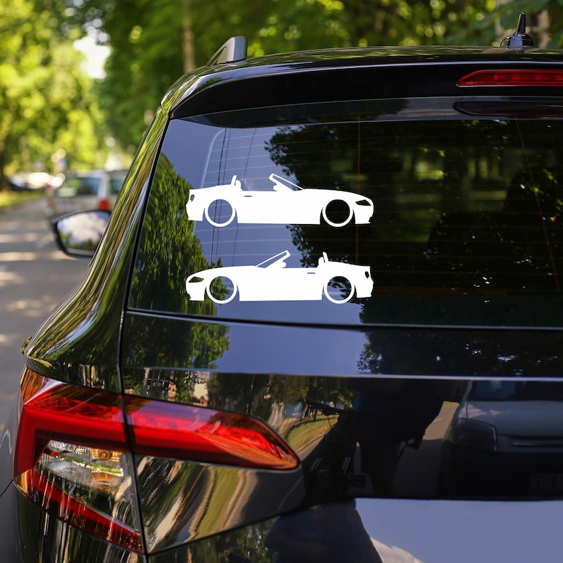 May include: Two white car decals of a convertible sports car are displayed on a black car window. The decals are side profiles, showing the car's body, wheels, and open top. The background is a blurred view of trees and other cars.