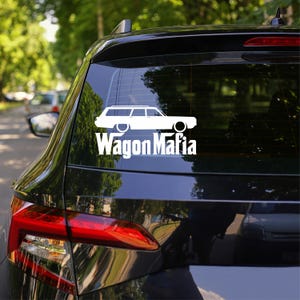 May include: White decal of a station wagon with the text "Wagon Mafia" on the rear window of a black car. The car is parked outdoors with trees visible in the background.