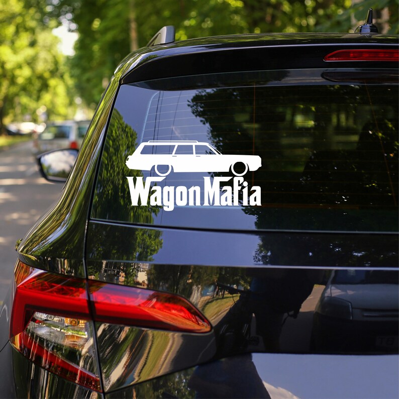 May include: White decal of a station wagon with the text "Wagon Mafia" on the rear window of a black car. The car is parked outdoors with trees visible in the background.