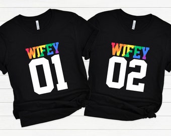 May include: Two black t-shirts with the text "WIFEY" in rainbow colors and the numbers "01" and "02" in white.