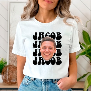 May include: White t-shirt with a black graphic design featuring the name "Jacob" in a retro, wavy font. The graphic is repeated multiple times and a person's face is centered in the design.