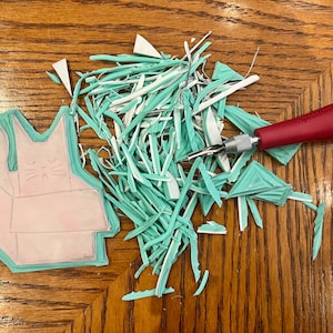 Może przedstawiać: Jasnoróżowa gumowa pieczątka z rysunkiem kota siedzącego w pudełku. Pieczątka jest otoczona jasnoniebieskimi i białymi wiórkami gumy. Na zdjęciu widoczne jest również czerwone i srebrne narzędzie do rzeźbienia.