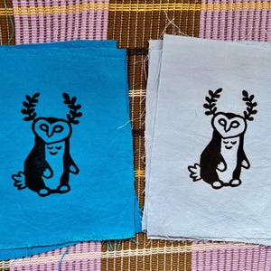 Pode incluir: Dois quadrados de tecido, um azul-turquesa e outro azul claro, cada um com uma ilustração preta de uma coruja abraçando uma figura menor. A coruja tem uma coroa de folhas. As bordas do tecido não estão acabadas.