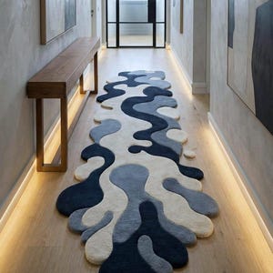 May include: A modern, abstract area rug in a hallway. The rug features a flowing, organic design with shades of navy, gray, and off-white. The rug's shape and color palette complement the light wood flooring and neutral walls.