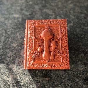 Könnte beinhalten: Ein kupferfarbenes Buch mit einem verzierten Design und einer 3D-Pilzskulptur auf dem Einband.