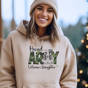 May include: A tan hoodie with the words "Proud ARMY Veteran's Daughter" printed on the front. The word "ARMY" is in a camouflage pattern. The hoodie has a front pocket and a drawstring hood.