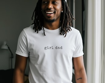May include: White t-shirt with the text "girl dad" in a simple, black font. The shirt is being worn by a person with dreadlocks and a white baseball cap. The person is smiling.