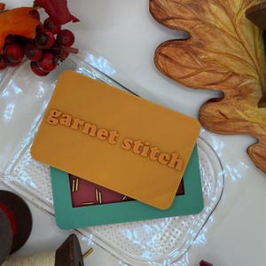 May include: A rectangular mustard yellow plastic card with the embossed text "garnet stitch" layered over a teal rectangular frame containing a dark red insert with small gold-colored metal pins arranged in a grid pattern.