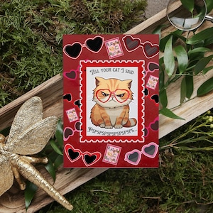 May include: A red greeting card featuring a cartoon cat wearing heart-shaped glasses and the text "Tell your cat I said". The card is decorated with heart-shaped sunglasses and postage stamp designs.
