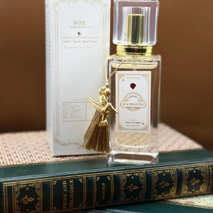 May include: A perfume bottle with a gold cap and a clear glass body, accompanied by a matching box. The bottle has a gold tassel with a hummingbird charm. The box and bottle are labeled "NOX BIBLIOTECA". The items rest on green books.
