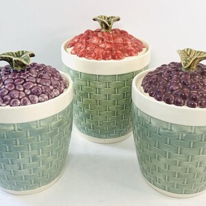 May include: Three ceramic jars with lids. The lids are shaped like clusters of grapes and are red, purple, and dark purple. The jars have a basket weave pattern in green and blue-green. The rims are white, and the lid handles are green.
