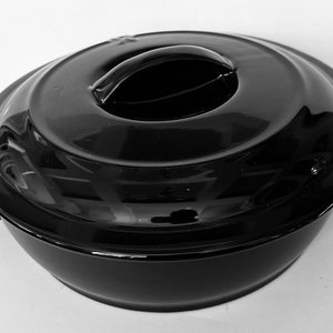 Könnte beinhalten: Eine schwarze, glänzende Auflaufform mit passendem Deckel. Der Deckel hat einen abgerundeten Griff zum einfachen Anheben. Die Schüssel hat zwei seitliche Griffe zum Tragen. Die Gesamtform ist rund mit einem leicht ausgestellten Boden.