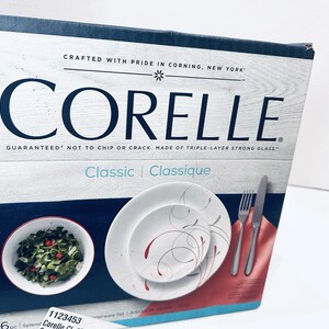 May include: A Corelle dinnerware set box with a white and blue design. The box features the Corelle logo and text that reads "Crafted with Pride in Corning, New York." The box also shows a plate, bowl, fork, and knife.