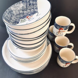 May include: A stack of white and blue ceramic plates and bowls with a snowman design. The plates and bowls have a blue rim and a blue and white pattern. There are also matching mugs with the same snowman design.