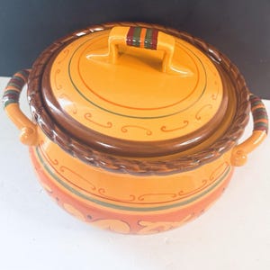May include: A round, lidded ceramic casserole dish in shades of orange, brown, and green. The dish has a braided rim and handles, with decorative lines and patterns. The lid features a handle with green and red stripes.