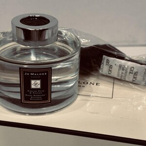 May include: A clear glass Jo Malone London diffuser with a silver cap, filled with liquid. The label reads "English Pear & Freesia". It sits on a white box with the brand name. A packet of silica gel is next to it.