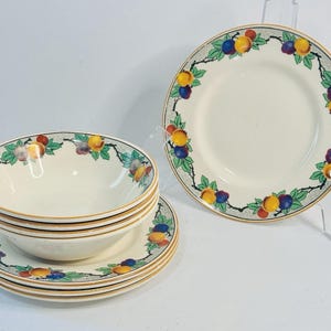 May include: A set of cream-colored ceramic dishes featuring a fruit design. The set includes bowls and plates, each adorned with a border of colorful fruits and green leaves. The rims have a gold trim, adding a touch of elegance to the tableware.
