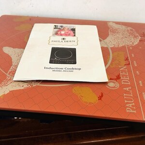 May include: A red Paula Deen induction cooktop with a black power cord. The cooktop has a patterned surface with the Paula Deen logo and a white instruction manual on top. The manual reads "Induction Cooktop".