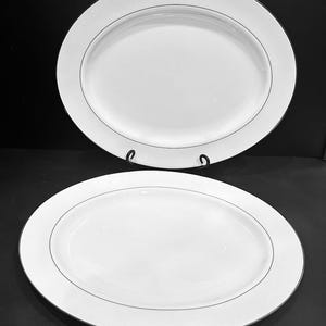 May include: Two white oval serving platters with a silver-colored rim and a subtle striped pattern. The platters are displayed on a black surface, showcasing their elegant design. Ideal for serving appetizers or main courses.