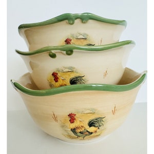 May include: Set of three ceramic bowls with a rooster design. The bowls are cream colored with a green rim and feature a rooster illustration with brown, yellow, and red accents.