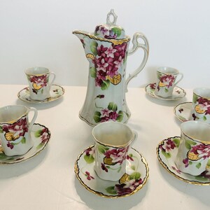 May include: A vintage floral tea set with a white teapot, six cups, and saucers. The set features a delicate floral pattern with pink and purple flowers and gold accents.