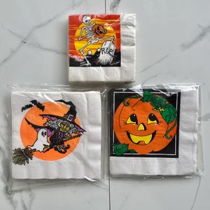 Puede incluir: Tres paquetes de servilletas de papel con temática de Halloween. Uno presenta un esqueleto sosteniendo una calabaza con una lápida que dice "RIP". Otro muestra una bruja en una escoba con un fantasma. El último tiene una calabaza sonriente.