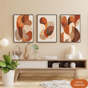 May include: Three framed abstract art prints. Each print features a unique design with organic shapes in shades of brown, orange, and beige against a cream background. The prints are displayed above a wooden console table with decorative items.